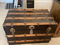 Front view of the antique travel trunk showing black body, wooden slats, brass clasps, and central lock.