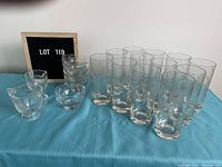 Full lot showing glassware including 12 tall drink glasses, 4 small parfait glasses, cream pitcher, and sugar bowl on blue cloth