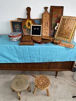 Wide view of the folk art lot showing multiple wooden plaques, boxes, and two small stools arranged on a surface.