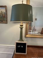 Green table lamp with drum shade and brass detailing shown on wooden surface against a background with paintings and mirror.