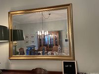 Large rectangular wall mirror with a gold-colored frame. The mirror reflects a dining room setting with furniture and lighting fixtures.