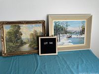 Two framed landscape paintings side by side on a blue surface with lot number board in between