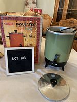 Full view of vintage Empire party coffee maker in green color next to original box and black sign with lot number. Lid placed in foreground showing black knob handle and stainless steel material.