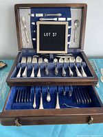 Original wooden flatware case opened showing two tiers lined with blue velvet holding silver plate flatware items in Remembrance pattern.