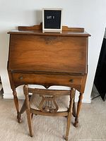 Full view of vintage wooden secretary desk with closed fold-down front and matching chair in front.