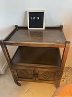 Front view of vintage wooden side table showing top surface, middle shelf, and double-door cupboard with carved panels and original round pulls. Curved legs with wheels are visible.