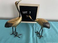 Both brass bird sculptures displayed on a blue cloth with a sign reading LOT 7 behind them.