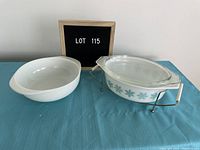 Photo showing both Pyrex dishes side by side with a lot number board behind, highlighting the plain white round dish and the teal snowflake patterned oval dish with lid on metal stand.