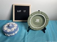 Photo of blue and white trinket box and green decorative plate on black stand, with lot sign in background.
