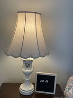 Full view of one white ceramic table lamp lit with white empire shade and letter board showing lot number