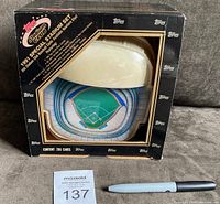 Outer cardboard box with window showing plastic stadium model inside. Box has wear and creases on edges, the window displays the stadium and card pack inside.
