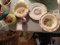Photo showing the Bunnykins serving pieces including two bowls and one plate, two small Bunnykins figurines, and the large Schmidt music box figurine on a table with auction tags.