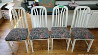 Four white wooden chairs with floral upholstered seats arranged in a row on hardwood floor.
