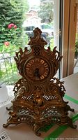 Front view of the vintage ornate metal table clock showing the clock face with Roman numerals and decorative gold-tone metalwork.