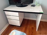 Angle showing front and left side of the white desk with three drawers and black trim.