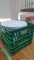 Stack of white Dudson Duraline dinner plates and serving bowl inside a green plastic milk crate, on wooden floor