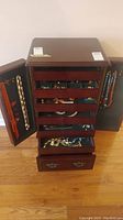 Wooden jewelry cabinet with doors open showing necklaces hanging and drawers with jewelry inside.