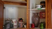 Photo showing large brown vase surrounded by mugs and cups, flower figurine, small candles, and shakers inside a wooden cabinet.