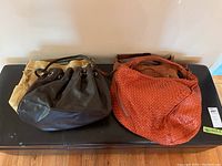 Four purses displayed on a black surface showing different colors and styles including beige fabric purse, orange woven leather hobo, and dark brown leather purse.