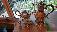 Side angled view of a silver plated tea set including teapot, coffee pot, sugar bowl with lid, creamer, and spoon on a small display stand.