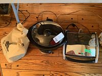 Photo showing panini press, brass slow cooker with glass lid, and Magic Bullet set in bag on wooden floor