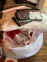Photo showing various folded table and kitchen linens stacked on a plastic bag, including a light-colored damask tablecloth and folded Christmas-themed cloth with green and red border and holly berry embroidery.