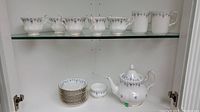Photo showing 7 teacups, 2 mugs on right, stacked saucers below on shelf, and teapot with lid on bottom shelf
