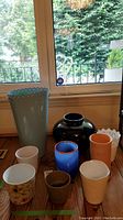Photo showing a collection of ceramic and glass vases and pots arranged on a wooden floor near a window. Includes a tall pale blue vase, a large black glossy vase, several ceramic pots in various colors and patterns, and a white woven wine cooler basket.