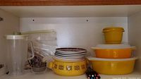 Top shelf with two yellow Pyrex baking dishes with lids stacked, yellow ceramic fondue pot with floral square design, nine fondue forks, and other glassware items
