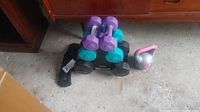 Photo showing stack of dumbbells, one kettlebell, ankle weights, and dumbbell stand on floor.