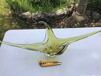 Top-down view showing the full elongated arms of the yellow glass sculpture placed on a white table outdoors with a utility knife for scale