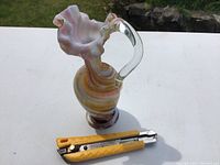 Full view of vintage art glass vase showing multi-colored swirl body, clear handle, and ruffled rim. Yellow utility knife placed next to it for size reference.