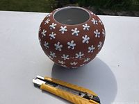 Overview of spherical pottery vase with white flower decorations and blue centers, showing shape and size next to utility knife for scale.