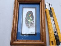 Framed feather art showing the full piece alongside a utility knife for scale.