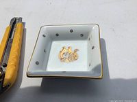 Small square white porcelain dish with gold trim and central gold decorative design, sitting on white surface