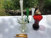 Five vintage colored glass vases displayed on white table outside. Clear ruffled vase in front, white tall vase, green tall vase, frosted white vase, and red scalloped vase grouped together.