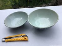Two antique glazed pottery bowls with blue scroll designs photographed on white surface next to a yellow cutting tool for scale.