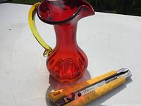 Red art glass jug with yellow handle viewed from the side showing spout and handle details alongside a box cutter for scale.