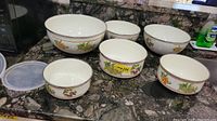 Set of six metal mixing bowls with lids on kitchen counter showing fruit motif decoration.