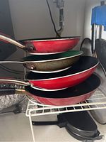 Four frying pans stacked on a wire rack inside a cabinet, showing red, green, black, and brown colors and worn condition