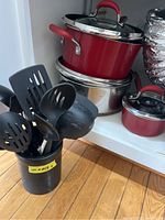 View of red cooking pots stacked together and black utensil holder with cooking utensils on wooden floor inside cabinet