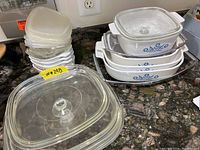 Overview on countertop: large clear Pyrex casserole with knob lid front left, flat glass lids and opaque plastic lids stacked behind, white CorningWare Cornflower dishes stacked right