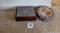 Wooden box with a decorative silver metal floral lid next to a round metal container with a colorful pattern.