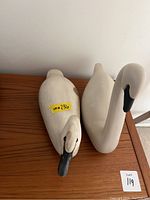 Two white swan figurines side by side on a wooden surface, viewed from above showing the body and necks.