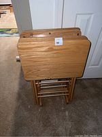 Front view of the wooden tray tables stacked on the stand showing the rectangular foldable tops and wooden legs folded beneath.