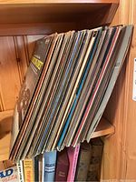 Side view of vinyl record albums stacked vertically on a wooden shelf, varying album cover designs visible, showing collection size and condition.