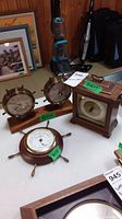 Three barometers displayed on white surface: two brass mounted on wooden base, one square wooden case, and one ship's wheel style barometer.