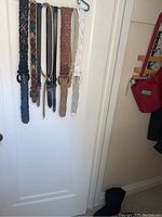 Photo of six women's belts hanging on a white door in different colors and styles including black braided, brown leather, and white perforated belts.