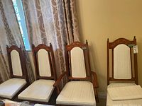 Front view of four wooden dining chairs with cream upholstered seat and backrest, positioned side by side. Two chairs on right have armrests (captain's chairs). Carved wood detail on top of backrest visible.