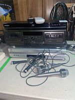Stack of electronic devices including Samsung Blu-ray player on the top, Technics stereo cassette deck in the middle, JVC DVD/CD player on the bottom, along with four wired microphones placed in front.